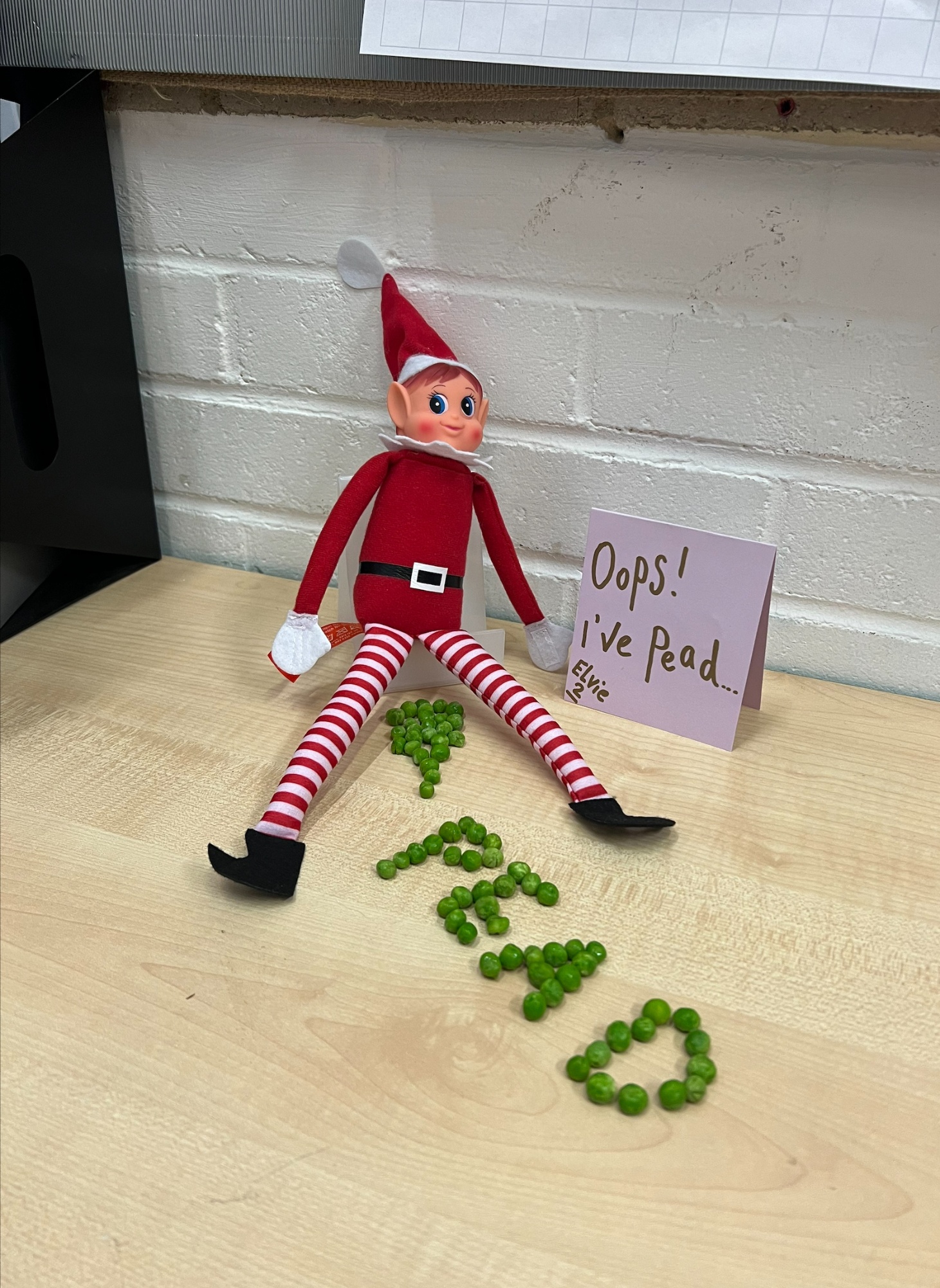 An elf sits with the word "PEAD" spelt in peas between it's legs.